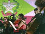 Orquestra tocando em frente a uma bandeira do estado de Santa Catarina