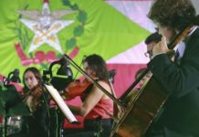 Orquestra tocando em frente a uma bandeira do estado de Santa Catarina