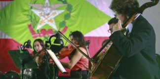 Orquestra tocando em frente a uma bandeira do estado de Santa Catarina