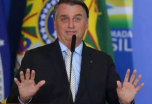 Bolsonaro sorrindo com as mãos para cima em um palanque, ele está de terno preto