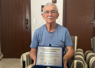 Professor zé sentado em uma poltrona com uma placa de homanenagem no colo dele