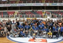 várias pessoas da equipe de basquetebol em cadeira de rodas joinville dentro do ginásio com a arquibancada de fundo