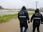 chuva em joinville, duas pessoas da defesa civil com capas de chuva, de costas, andando em uma rua de barro em joinville