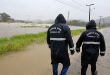 chuva em joinville, duas pessoas da defesa civil com capas de chuva, de costas, andando em uma rua de barro em joinville