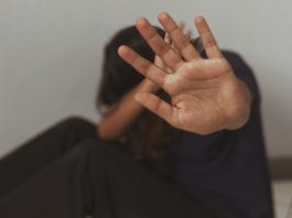 uma mulher sentada no chão de lado, com a cabeça baixa tentando se proteger com uma mão aberta esticada para frente e outra ao redor da cabeça