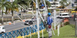 Dois homens um subindo uma escada que está encostada em um poste e o outro está no chão olhando para cima. Eles estão instalando luzes para o natal em Joinville