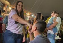 mulher em pé sorrindo com a mão dada com homem abaixado, de costas, pedindo ela em casamento em meio as luzes de natal na abertura de natal em joinviille