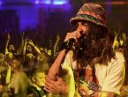Cantor Armandinho com os cabelos longos soltos e um chapeu na cabeça. ele canta para uma plateia