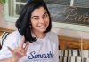 Mulher sorridente, vestindo uma camiseta branca com o logo da Semearhis, uma startup de Joinville que promove a empregabilidade de pessoas com deficiência, faz o gesto de 'eu te amo' em Libras. O fundo da imagem é composto por uma decoração de madeira com plantas e uma placa ao fundo.