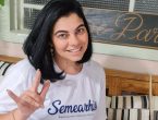 Mulher sorridente, vestindo uma camiseta branca com o logo da Semearhis, uma startup de Joinville que promove a empregabilidade de pessoas com deficiência, faz o gesto de 'eu te amo' em Libras. O fundo da imagem é composto por uma decoração de madeira com plantas e uma placa ao fundo.