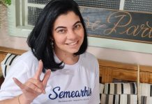 Mulher sorridente, vestindo uma camiseta branca com o logo da Semearhis, uma startup de Joinville que promove a empregabilidade de pessoas com deficiência, faz o gesto de 'eu te amo' em Libras. O fundo da imagem é composto por uma decoração de madeira com plantas e uma placa ao fundo.